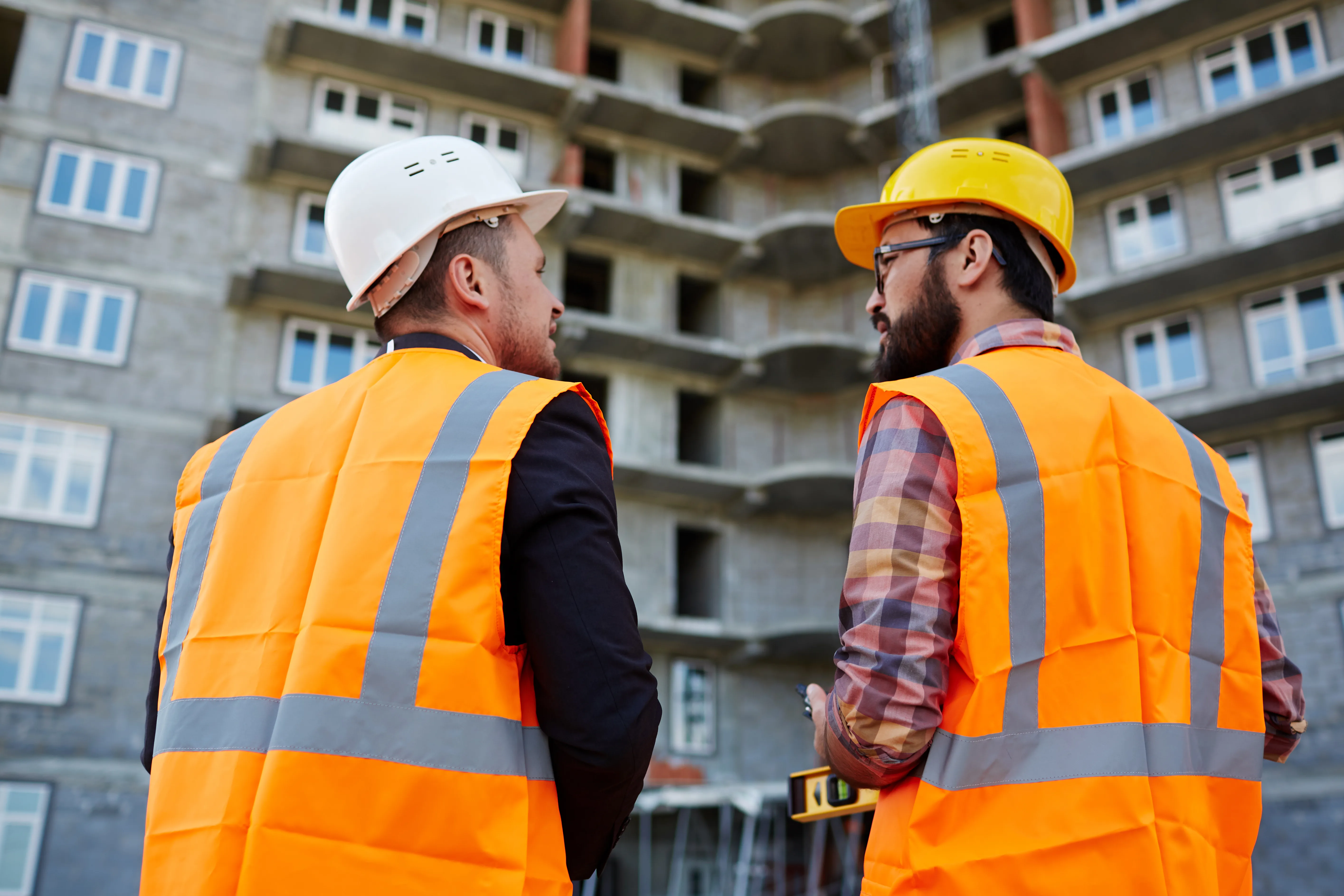 Backs of two builders in uniform having discussion on background of unfinished edifice
